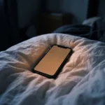 Phone lying face up on a bed sheet at night, its blank screen glowing warm amber in the dark.