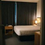 Dimly lit hotel room at night with a made bed and curtains drawn, lit only by a low warm lamp.