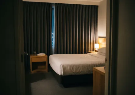Dimly lit hotel room at night with a made bed and curtains drawn, lit only by a low warm lamp.