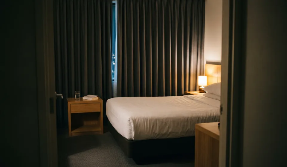 Dimly lit hotel room at night with a made bed and curtains drawn, lit only by a low warm lamp.