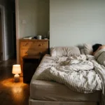 An empty side of a bed with the covers lifted, a sleeper's shape on the other side, and a small floor lamp lighting the floor toward a door left ajar.