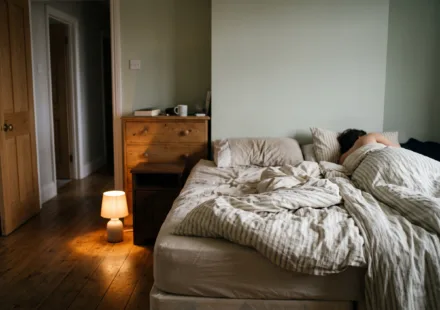 An empty side of a bed with the covers lifted, a sleeper's shape on the other side, and a small floor lamp lighting the floor toward a door left ajar.