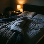 A bed in a dark room with the duvet bunched to one side and the other half bare