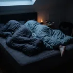 A double bed in a dim bedroom with one duvet pulled high on one side and a bare foot sticking out on the other.