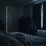 View from a pillow across a dark bedroom, with a thin amber line of light under the door and a pale seam between the curtains