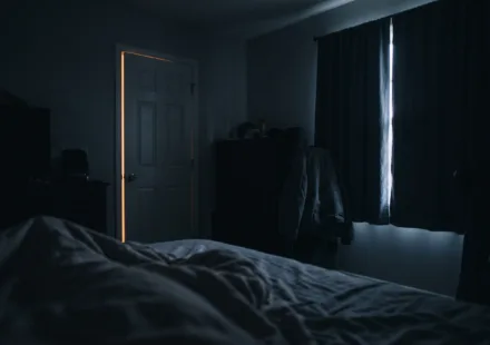 View from a pillow across a dark bedroom, with a thin amber line of light under the door and a pale seam between the curtains
