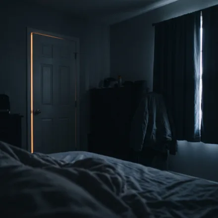 View from a pillow across a dark bedroom, with a thin amber line of light under the door and a pale seam between the curtains