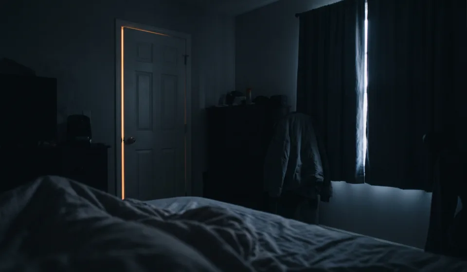 The Bedroom Audit: Eleven Minutes, Lights Off, From the Pillow View from a pillow across a dark bedroom, with a thin amber line of light under the door and a pale seam between the curtains