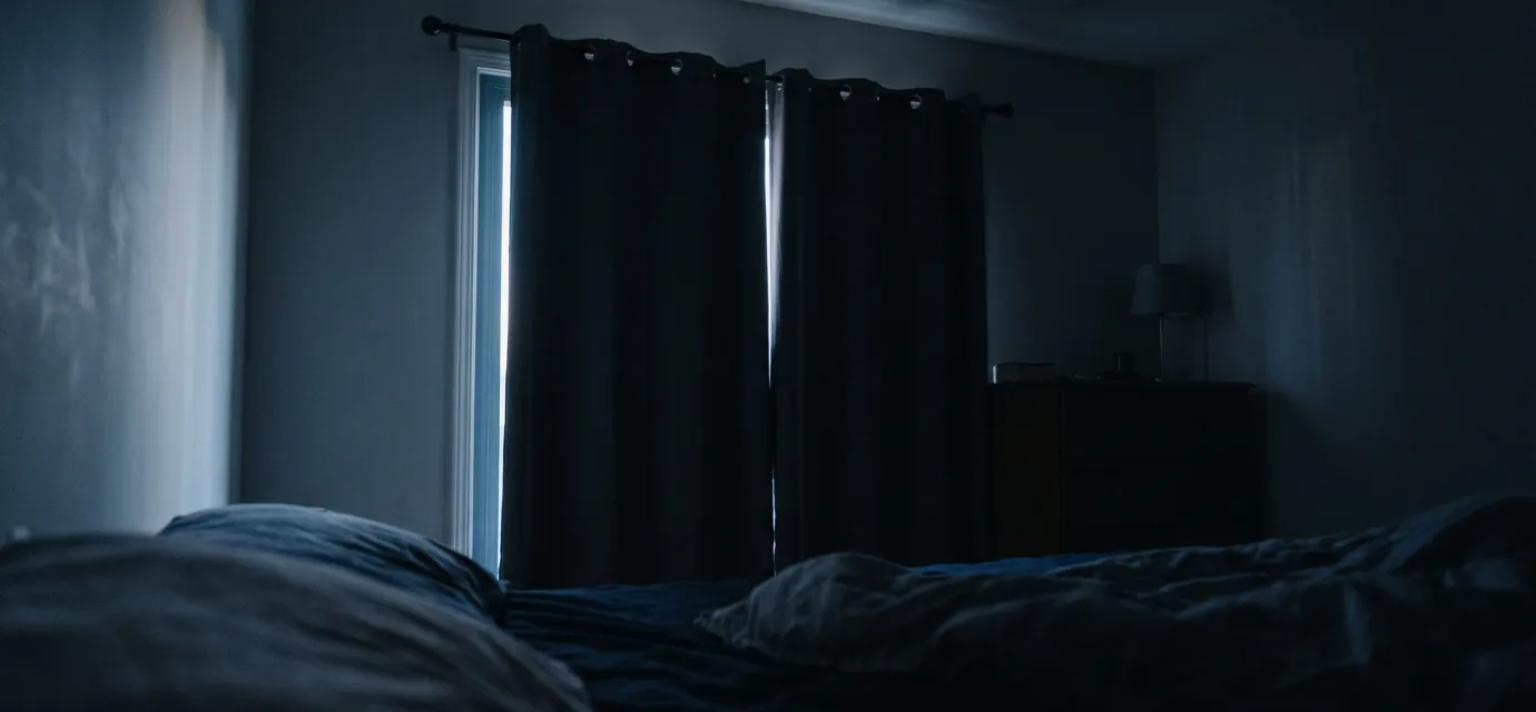 A dark bedroom just before sunrise, with a thin blade of pale light slipping along the edge of closed curtains and stretching across the ceiling