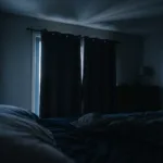A dark bedroom just before sunrise, with a thin blade of pale light slipping along the edge of closed curtains and stretching across the ceiling