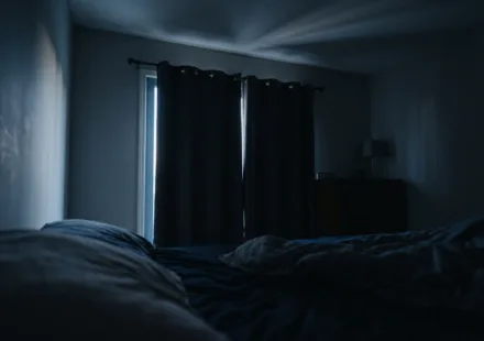 A dark bedroom just before sunrise, with a thin blade of pale light slipping along the edge of closed curtains and stretching across the ceiling