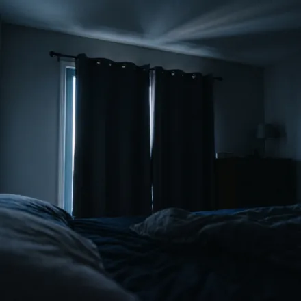A dark bedroom just before sunrise, with a thin blade of pale light slipping along the edge of closed curtains and stretching across the ceiling